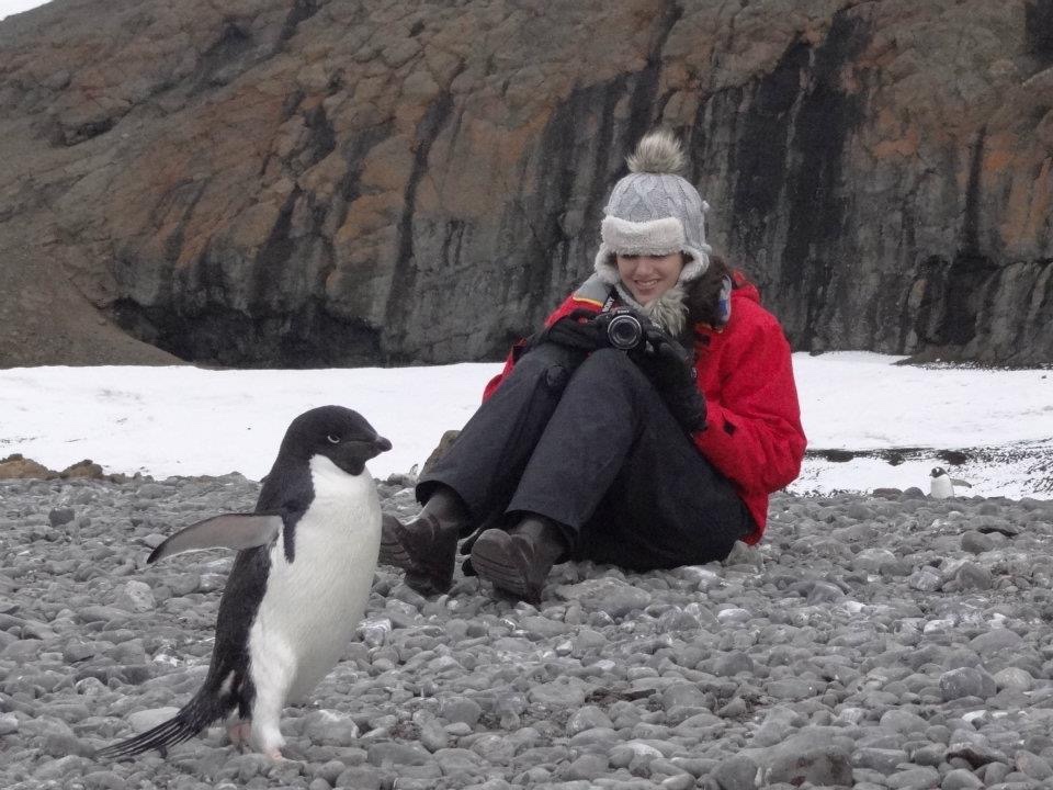 Woman wearing a red jacket and grey hat, sitting on rocks taking a photo of a adeloo penguin in Antarctica