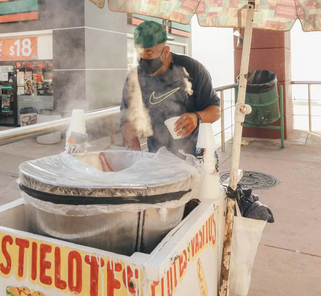 Esquites vendor on the street holding a cup to fill from the steaming metal pot in front of him