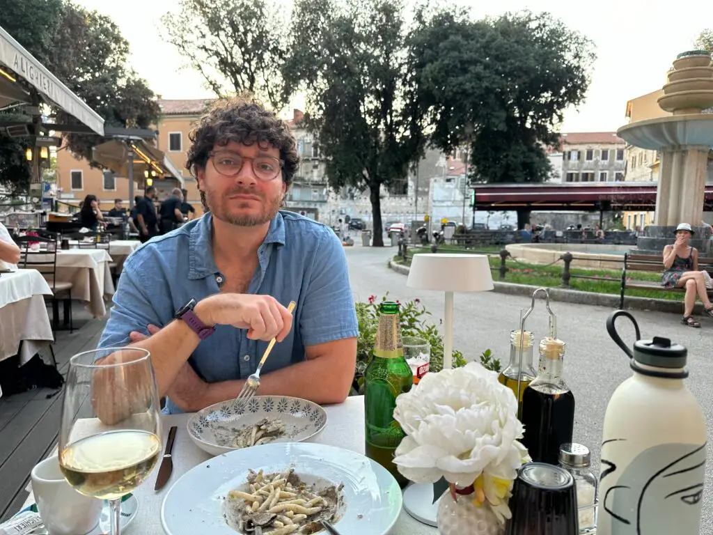 Curly haired man in a blue linen shirt eating Croatian pasta with truffles in a plaza in Pula as it’s pone of the best things to do in Pula