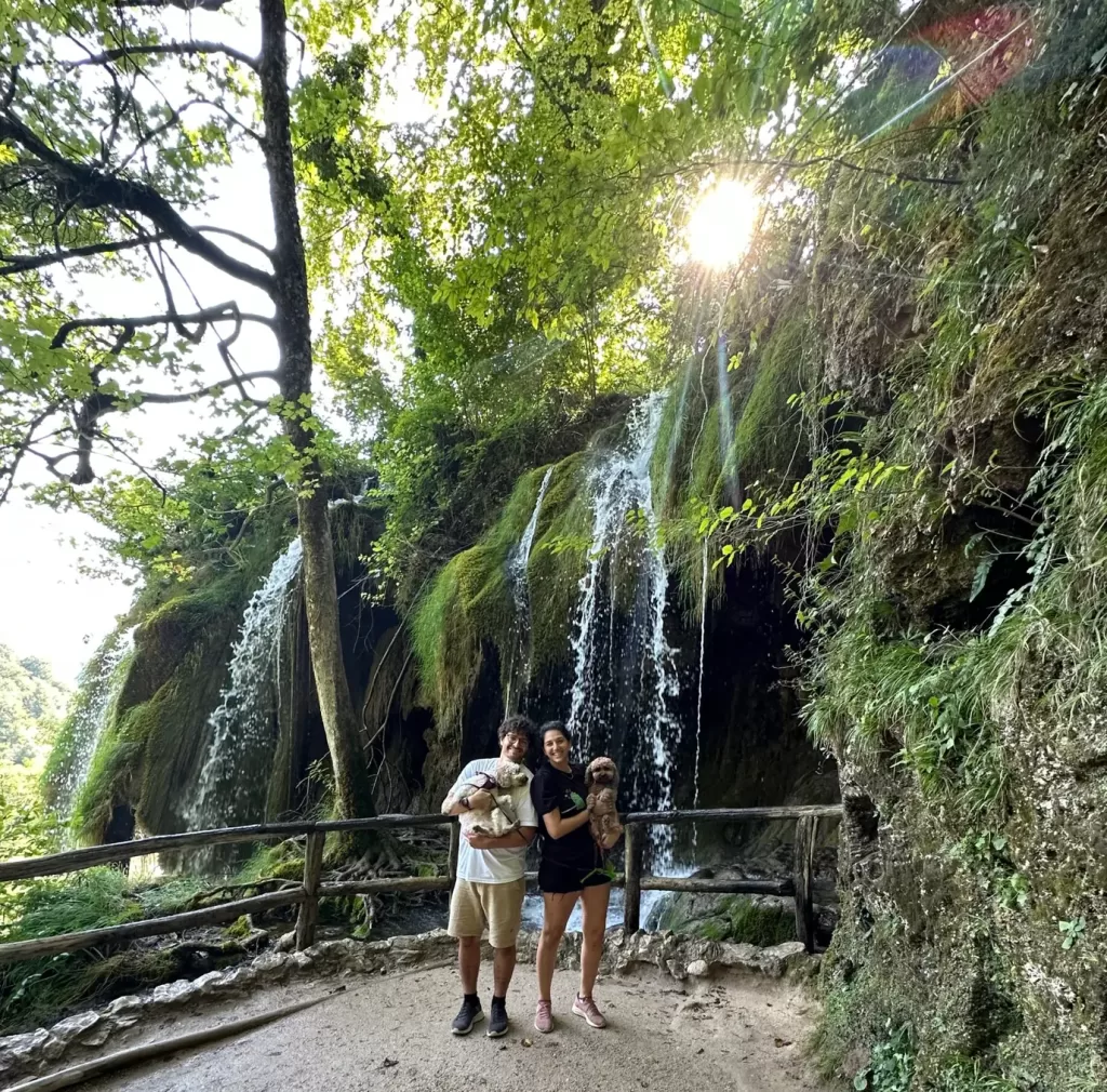 Man and woman each holding a dog in front of a waterfall with lush green vegetation in the background at Plitvice Lakes National Park