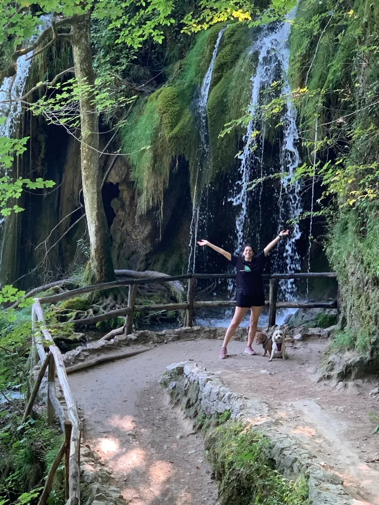 Woman in all black posing with arms up and open with two dogs by her feet in front of Veliki Prštavac Waterfall in Plitvice Lakes